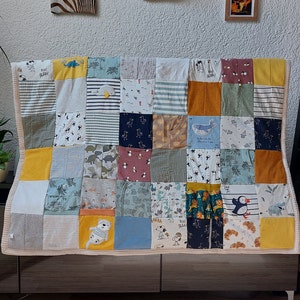 Può includere: Una trapunta patchwork realizzata con vari capi di abbigliamento per bambini. La trapunta presenta una varietà di colori e motivi, tra cui strisce, dinosauri e personaggi dei cartoni animati. La trapunta è esposta su un mobile marrone scuro.