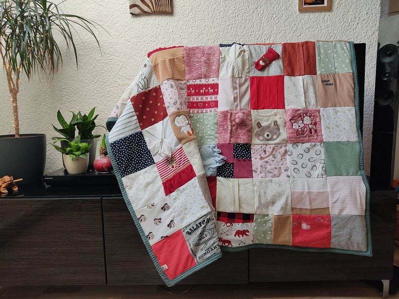 Puede incluir: Un edred&oacute;n de retazos hecho de varias telas, incluyendo ropa de beb&eacute;. El edred&oacute;n presenta cuadrados de diferentes colores y patrones, como lunares, rayas y dise&ntilde;os de animales. Los bordes est&aacute;n ribeteados con un borde turquesa.