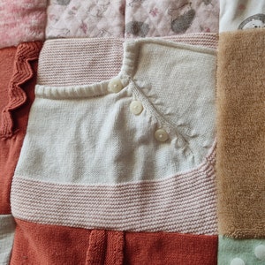 Puede incluir: Un edred&oacute;n patchwork hecho con ropa de beb&eacute;. El edred&oacute;n presenta un c&aacute;rdigan blanco tejido con botones, una secci&oacute;n a rayas rosas y blancas y secciones con estampados de conejos y erizos. El tema es beb&eacute; y guarder&iacute;a.