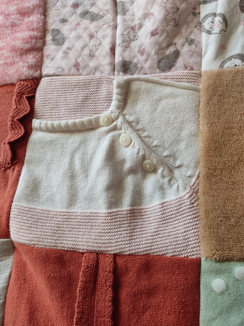 Puede incluir: Un edred&oacute;n patchwork hecho con ropa de beb&eacute;. El edred&oacute;n presenta un c&aacute;rdigan blanco tejido con botones, una secci&oacute;n a rayas rosas y blancas y secciones con estampados de conejos y erizos. El tema es beb&eacute; y guarder&iacute;a.