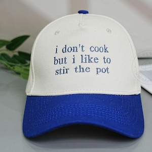 以下が含まれることがあります： 「i don't cook but i like to stir the pot」の刺繍が入った白と青の野球帽。 つばは青色で、上部に青色のボタンが付いています。 帽子は灰色の表面にあります。