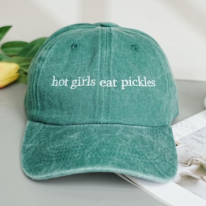 Op de afbeelding: Een groene baseballcap met de tekst "hot girls eat pickles" in het wit geborduurd. De cap heeft een gebogen klep en een licht vervaagde uitstraling, wat wijst op een vintage of versleten look. De cap is gemaakt van een zachte stof.