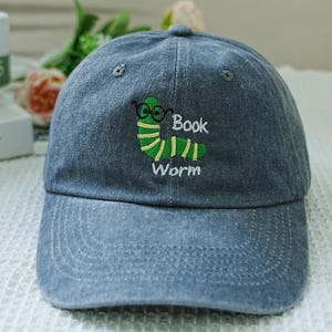 Puede incluir: Una gorra de béisbol de denim azul con un gusano de libro bordado en verde que lleva gafas y el texto "Book Worm".