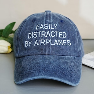 Może przedstawiać: Niebieska dżinsowa czapka z daszkiem z białym haftem "EASILY DISTRACTED BY AIRPLANES". Czapka jest lekko wyblakła, co nadaje jej wygląd vintage. Klasyczny sześciopanelowy design.