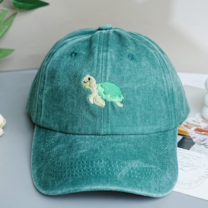 Pode incluir: Boné de beisebol turquesa com aba curva. O boné apresenta um desenho de tartaruga bordada em verde e bege na frente. O chapéu é feito de um tecido macio e texturizado.