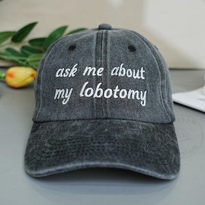 Peut inclure: Casquette de baseball grise avec le texte brodé "ask me about my lobotomy" en blanc. La casquette a une visière incurvée et un aspect vintage vieilli. C'est un accessoire décontracté.
