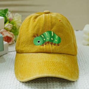 Puede incluir: Una gorra de béisbol amarilla con una oruga verde bordada en la parte delantera.