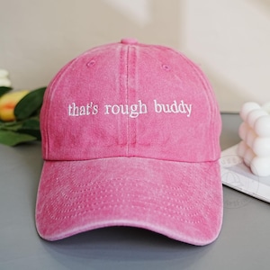 Pode incluir: Boné de beisebol rosa com o texto bordado "that's rough buddy" em branco. O boné tem uma aba curva e um design clássico de seis painéis. O tecido parece ser algodão lavado.
