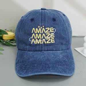 May include: A dark blue denim baseball cap featuring the word AMAZE embroidered three times in yellow thread, stacked vertically with a thin yellow orbital line encircling the text and three small star icons above the lettering.