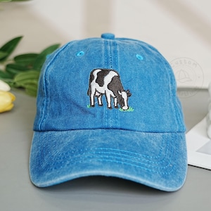 Może przedstawiać: Niebieska dżinsowa czapka z daszkiem z haftem przedstawiającym czarno-białą krowę z przodu. Czapka ma zakrzywiony daszek i guzik na górze. Krowa pasie się na zielonej trawie.