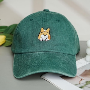 May include: Green baseball cap with a curved brim. The cap features an embroidered design of a corgi's rear end in shades of brown and white. The cap is made of a soft, textured fabric.