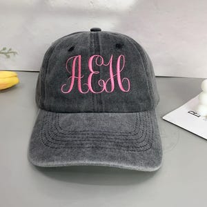Peut inclure: Une casquette de baseball gris anthracite avec une finition délavée et usée. Le panneau avant présente une broderie en fil rose vif formant les lettres AEH dans une police cursive fluide. La casquette possède une visière courbée et des coutures.