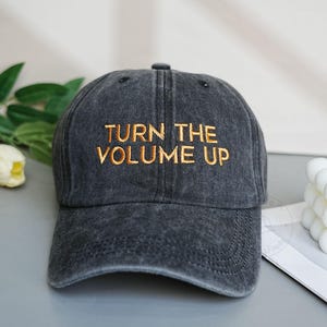 Könnte beinhalten: Dunkelgraue Baseballkappe mit dem goldfarbenen Schriftzug "TURN THE VOLUME UP". Die Kappe hat einen gebogenen Schirm und ein klassisches Sechs-Panel-Design. Der Hut ist aus einem weichen, abgenutzten Material.