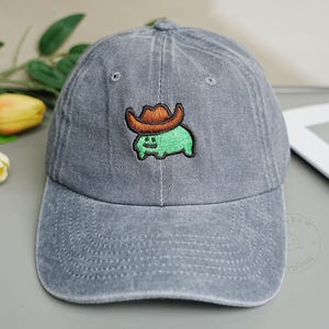 Puede incluir: Gorra de béisbol gris con visera curva. La gorra presenta una criatura verde bordada con un sombrero de vaquero marrón. El sombrero está hecho de un material suave y duradero, adecuado para uso casual.