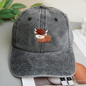 Puede incluir: Gorra de béisbol de mezclilla gris con visera curva. La gorra presenta un diseño bordado de una vaca de las Tierras Altas marrón y blanca. La gorra tiene un aspecto desgastado y una corona redondeada.