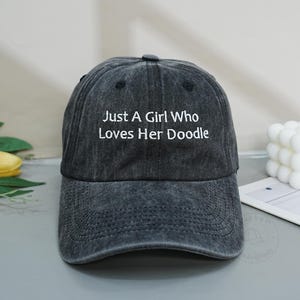 Könnte beinhalten: Eine anthrazitfarbene Baseballkappe mit verwaschener Optik. Auf der Vorderseite befindet sich ein weißer Stickschriftzug mit dem Text, Just A Girl Who Loves Her Doodle, der mittig in einer serifenlosen Schrift über dem gebogenen Schirm platziert ist.