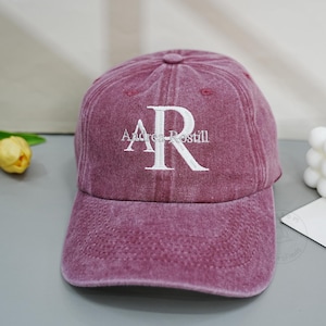 Op de afbeelding: Een kastanjebruine baseballpet met een verweerde, verwassen textuur. De voorkant is voorzien van wit geborduurde tekst "Andrea-Rostill" met een groot, gestileerd "AR"-monogram gecentreerd boven de naam in een klassiek schreeflettertype.