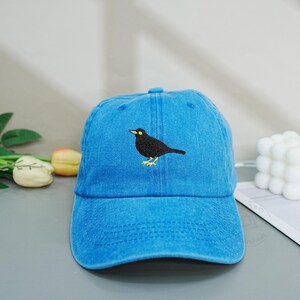 Könnte beinhalten: Eine leuchtend blaue Baseballkappe mit einer mittigen Stickerei eines schwarzen Vogels mit gelbem Schnabel und gelben Füßen. Die Kappe hat eine strukturierte Krone, einen gebogenen Schirm und sichtbare Nähte auf den vorderen Paneelen.
