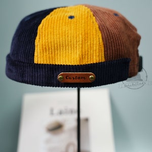 Puede incluir: Un gorro de pana azul, amarillo y marrón con un parche de cuero marrón que dice "Custom".