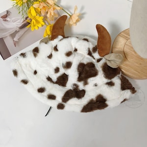 May include: A white and brown cow print bucket hat with brown fuzzy ears and horns. The hat has a black drawstring.