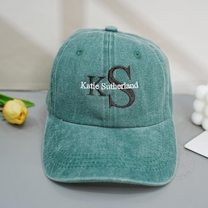 Pode incluir: Um boné de beisebol verde lavado com as letras K e S bordadas em preto, e o texto branco Katie Sutherland sobreposto no centro. O boné tem uma copa estruturada, uma aba curva e costuras verdes a condizer em todo o tecido.