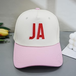 Op de afbeelding: Een baseballpet met een gebroken witte kroon en een lichtroze klep. Op de voorkant van de kroon staan de letters JA geborduurd in het rood met een lichtroze omlijning. De pet heeft een klein roze knoopje in het midden aan de bovenkant.