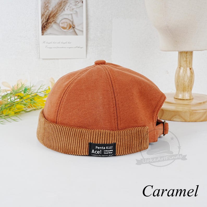 Może przedstawiać: Beanie w kolorze karmelowym z brzegiem z weluru i czarną naszywką z napisem "Penta Kill! Ace! God Tier" białymi literami.