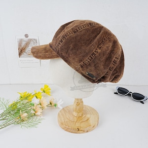 Puede incluir: Una gorra de estilo newsboy de pana marrón con una visera curvada y una visera pequeña y plana. La gorra está sobre un soporte de madera.