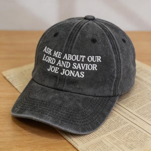 May include: A charcoal gray baseball cap with the embroidered text "ASK ME ABOUT OUR LORD AND SAVIOR JOE JONAS" in white. The cap has a curved brim and a button on top. The hat is sitting on a piece of newspaper.