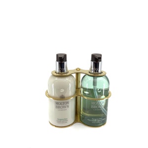 May include: A double bottle set by Molton Brown London, held in a brass-toned frame. One bottle contains hand lotion, the other hand wash. Both have dark brown pump dispensers. The bottles are labelled with product and brand details.
