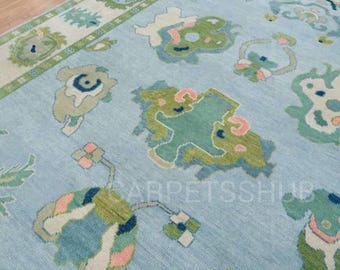 May include: A light blue area rug featuring a repeating pattern of abstract shapes in shades of green, cream, and soft pink. The rug displays the text CARPETSSHUB in a semi-transparent font across the center of the woven surface.