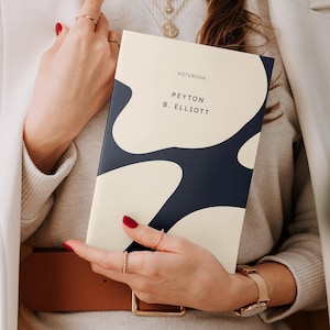 Könnte beinhalten: Eine Frau hält ein cremefarbenes und marineblaues Notizbuch mit dem Text "NOTEBOOK PEYTON B. ELLIOTT" auf dem Einband.