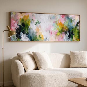 Könnte beinhalten: Ein großes rechteckiges Leinwandgemälde mit abstrakten Pinselstrichen in Rosa, Gelb, Waldgrün und Weiß. Das Kunstwerk ist in helles Holz gerahmt und hängt horizontal an einer Wand über einem cremefarbenen Sofa mit strukturierter Oberfläche.