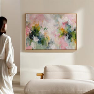 以下が含まれることがあります： 木製のフレームに入った抽象的な花の絵画。ピンク、白、緑、黄色の筆致が混ざり合い、柔らかくカラフルな構図を作り出しています。絵画は、クリーム色のソファの上にある白い壁に展示されています。