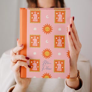 Può includere: Un'agenda rosa con dorso arancione e copertina con un motivo ripetuto di soli, lune, stelle e illustrazioni incorniciate di una statua. Le parole "Mia's Planner" sono scritte in corsivo.