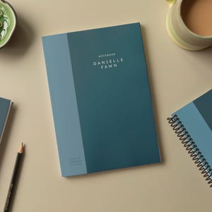 Op de afbeelding: Drie blauwe notitieboekjes met de tekst "NOTEBOOK DANIELLE FAWN" op de omslag. Twee notitieboekjes zijn gesloten en één is open met een spiraalbinding.