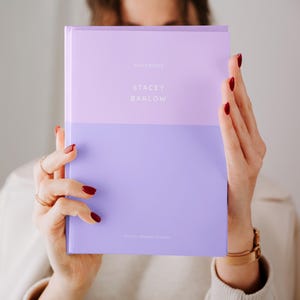 Könnte beinhalten: Ein lila Notizbuch mit dem Text "NOTEBOOK" und "STACEY BARLOW" in weißer Schrift. Das Notizbuch wird von einer Person mit rotem Nagellack gehalten.