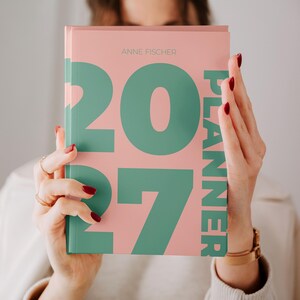 Könnte beinhalten: Ein Hardcover-Planer mit einem hellrosa Einband, der große, kräftige, blaugrüne Zahlen mit der Aufschrift 2027 und das vertikal in Blaugrün auf der rechten Seite gedruckte Wort PLANNER zeigt. Der Name ANNE FISCHER steht oben in kleinen, dünnen Buchstaben.