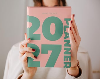 Könnte beinhalten: Ein Hardcover-Planer mit einem hellrosa Einband, der große, kräftige, blaugrüne Zahlen mit der Aufschrift 2027 und das vertikal in Blaugrün auf der rechten Seite gedruckte Wort PLANNER zeigt. Der Name ANNE FISCHER steht oben in kleinen, dünnen Buchstaben.