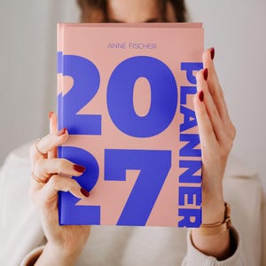 Peut inclure: Un agenda couleur pêche avec un grand texte bleu en gras indiquant 2027 et PLANNER. Le nom ANNE FISCHER est imprimé en plus petit texte bleu en haut au centre de la couverture. Le livre est tenu à la verticale, montrant clairement le design.
