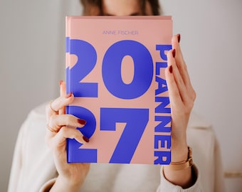 Könnte beinhalten: Ein pfirsichfarbener Planer mit großem, fettgedrucktem blauem Text, der 2027 und PLANNER lautet. Der Name ANNE FISCHER ist in kleinerem blauem Text oben mittig auf dem Vorderdeckel aufgedruckt. Das Buch wird aufrecht gehalten und zeigt das Design.