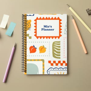 Może przedstawiać: Spiralny notatnik z kolorową, wzorzystą okładką z lampartem, brzoskwiniami i geometrycznymi kształtami. Notatnik oznaczony jest jako "Mia's Planner" i otoczony ołówkami, karteczkami samoprzylepnymi i spinaczem na neutralnej powierzchni.
