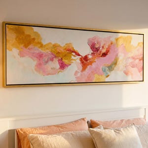 May include: Abstract painting with a gold frame. The artwork features a blend of pink, orange, and yellow hues, creating a soft, cloud-like effect. The painting is mounted above a white bed frame with peach-colored pillows.