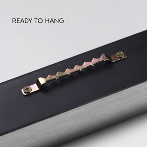 May include: Close-up of a black frame with a silver metal edge. A sawtooth hanger is attached to the back of the frame with a screw. The text "READY TO HANG" is visible at the top of the image.