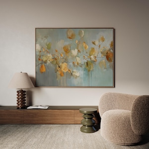 May include: Large framed abstract floral painting with a light blue and green background. The artwork features yellow and orange flower-like shapes on branches. The painting is displayed above a wooden cabinet, next to a lamp and a beige armchair.