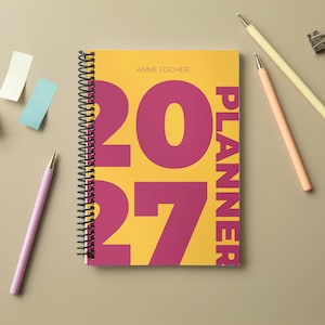 Könnte beinhalten: Ein spiralgebundener 2027-Planer mit gelbem Einband, großen magentafarbenen Ziffern und dem Text ANNE FISCHER PLANNER. Der Planer wird von drei Stiften in Helllila, Pfirsich und Creme, einer Metallklammer und drei Haftnotizen begleitet.