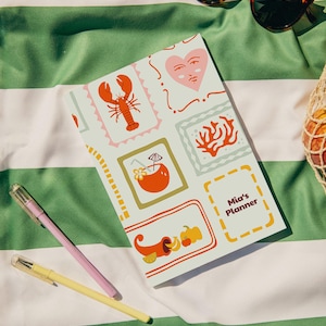 Può includere: Un'agenda con sfondo chiaro e illustrazioni colorate di un'aragosta, un cuore, coralli e un cocktail. L'agenda è etichettata "Mia's Planner" ed è accanto a una penna rosa e gialla e occhiali da sole su un asciugamano a righe verdi e bianche.