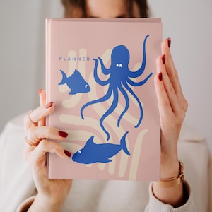 Op de afbeelding: Een roze planner met blauwe illustraties van een octopus, een vis en een haai. Het woord "PLANNER" staat bovenaan gedrukt. De planner wordt vastgehouden door een persoon met rode nagellak.