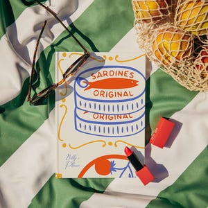Può includere: Scena sulla spiaggia con un libro con l'illustrazione di una scatoletta di sardine, occhiali da sole, un rossetto rosso e una borsa a rete con pesche. Il libro riporta la scritta "Sardines Original". La scena è su un asciugamano a righe verdi e bianche.