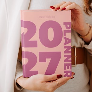 Peut inclure: Un agenda à couverture rigide rose clair avec le texte violet 2027 en grande taille et le mot PLANNER imprimé verticalement sur le côté droit. Le nom ANNE FISCHER est imprimé en petits caractères fins et clairs au centre supérieur.